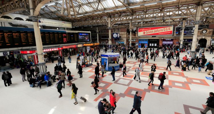 a busy concourse of a uk train station. The image shows people waiting for trains and the display board with the rail departures
